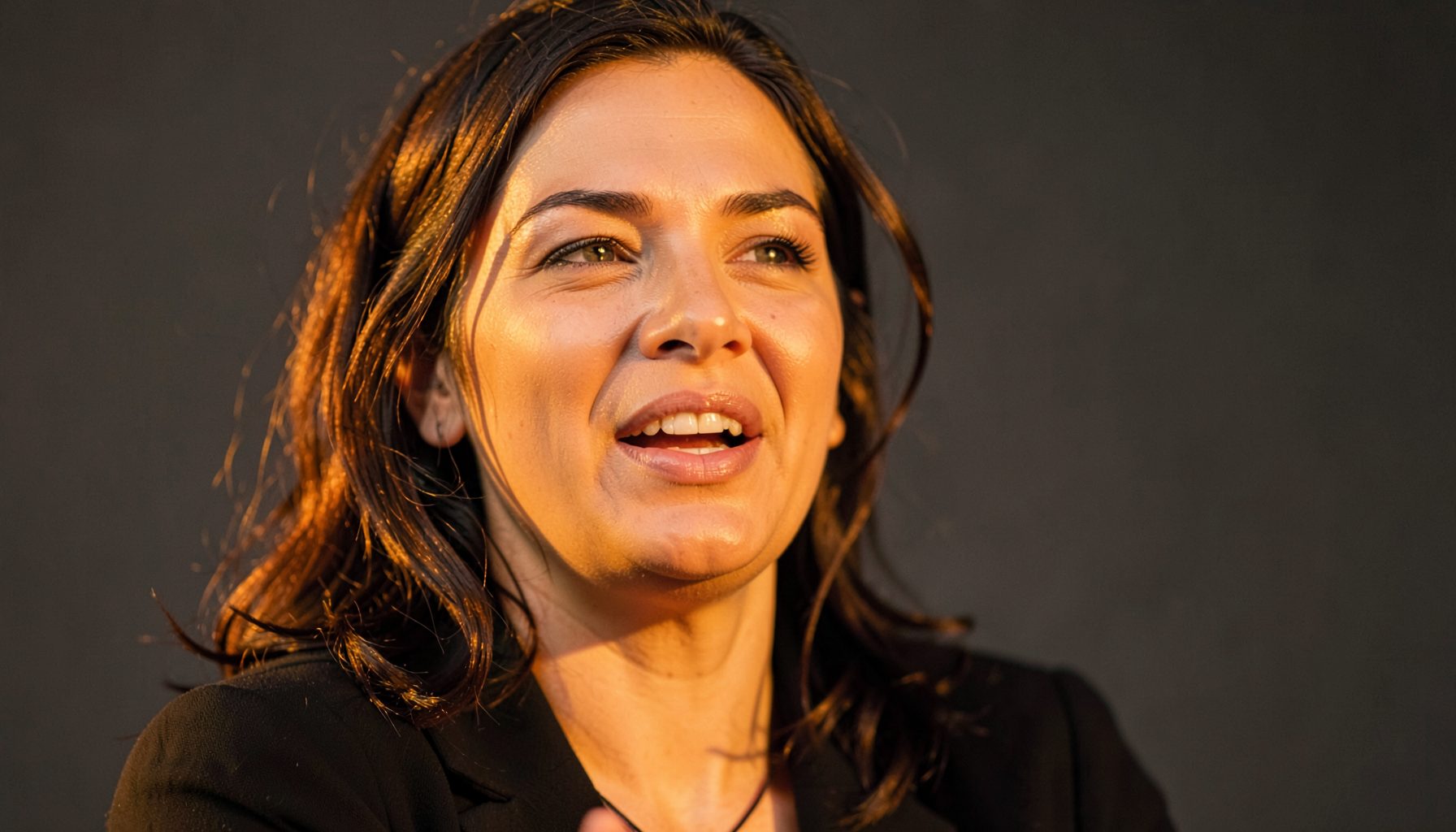 A close-up of a woman speaking passionately, bathed in warm golden light against a muted charcoal backdrop, conveying confidence and authenticity.
