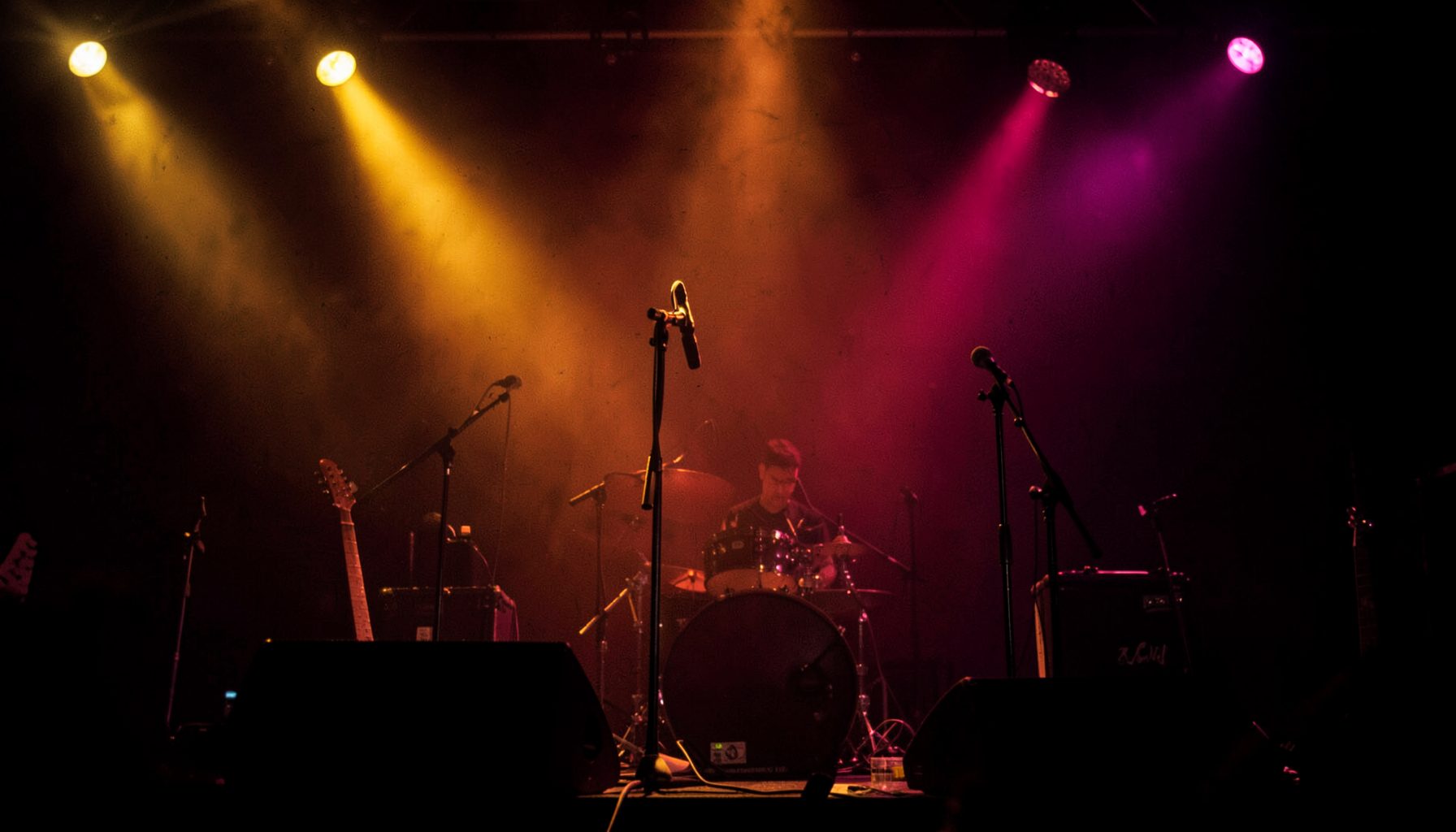 A dimly lit stage with warm amber and magenta spotlights casting a glow over microphone stands and instrument silhouettes, evoking an intimate live-music atmosphere.