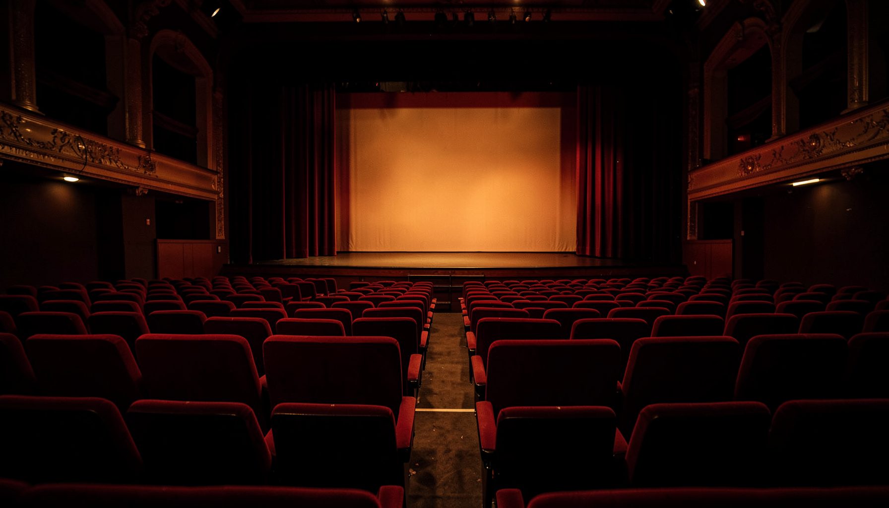 A dimly lit theater interior with deep red velvet seats and a glowing stage, warm amber and crimson tones filling the space