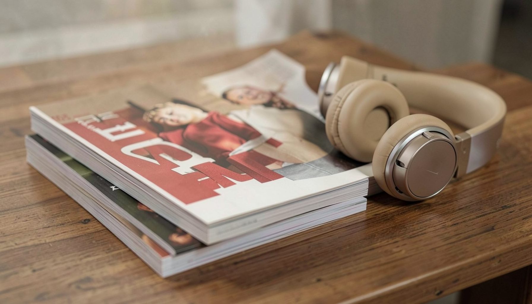 A stack of magazines and headphones on a wooden desk, muted earth tones with pops of red and cream