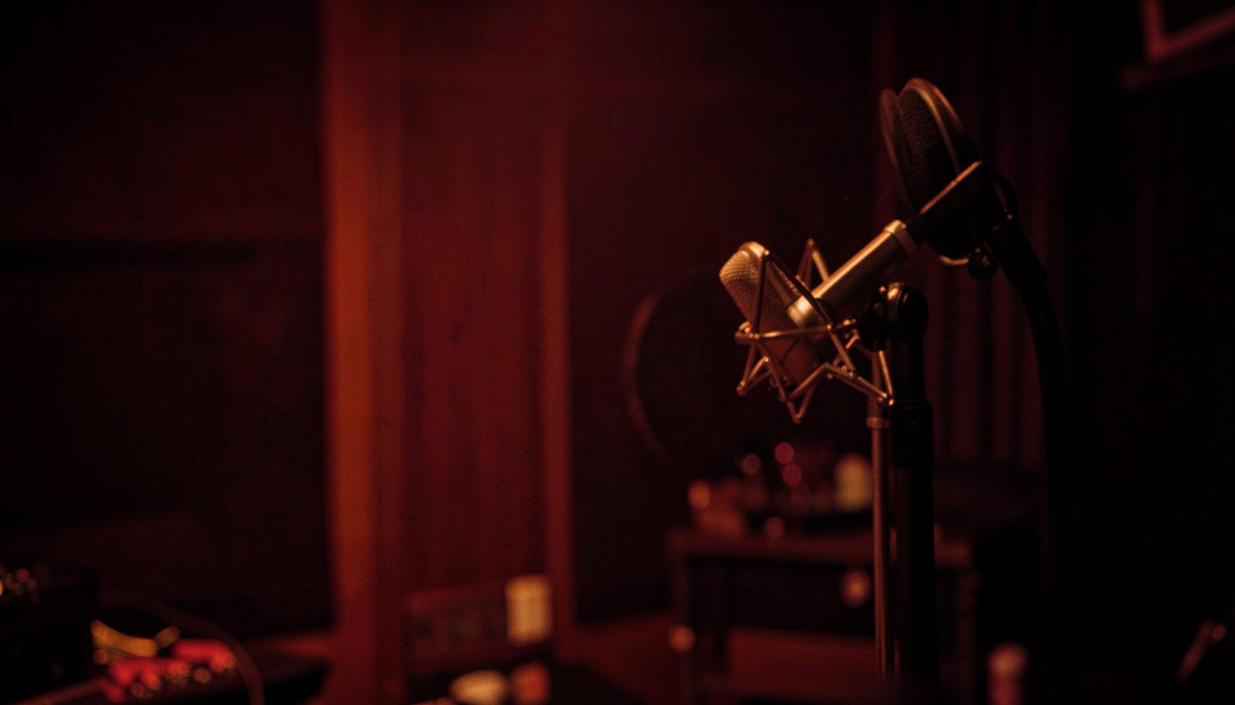 A warm, dimly lit recording studio with a microphone on a stand in the foreground, deep red and amber tones, moody and intimate atmosphere