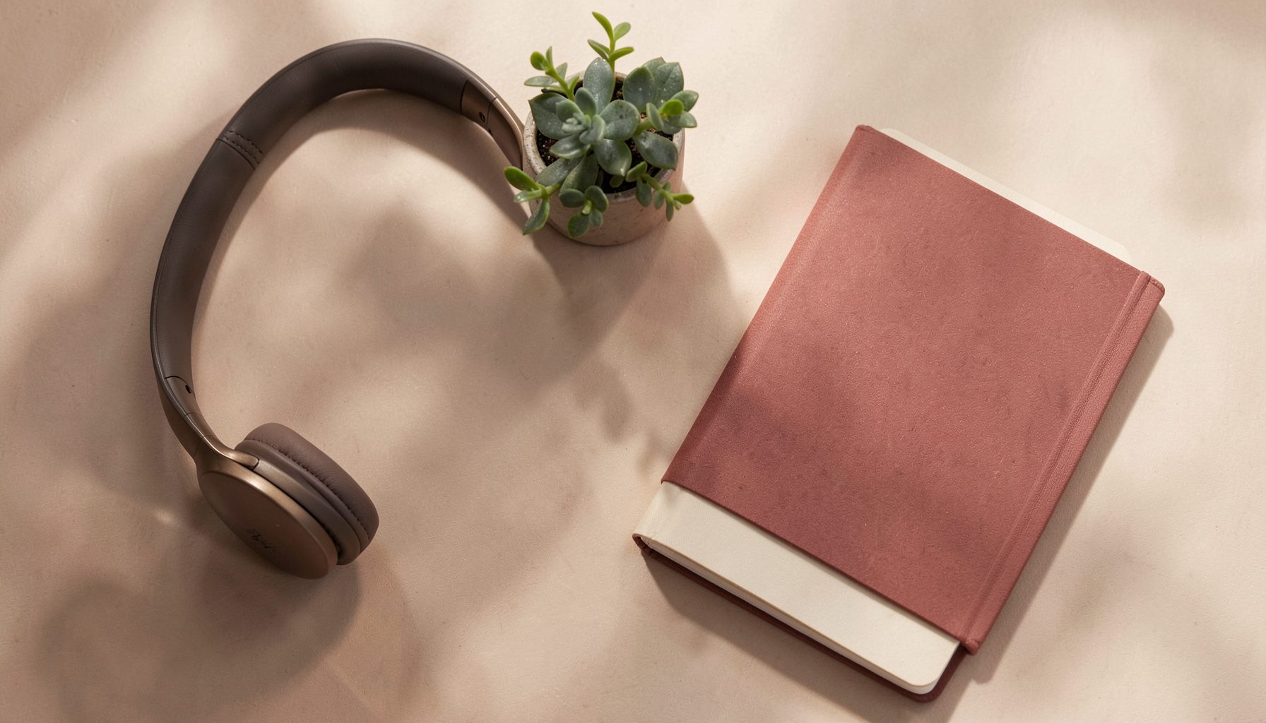 A warm, softly lit flat-lay of lifestyle items including headphones, a small plant, and a notebook in muted red, cream, and charcoal tones, evoking a relaxed editorial mood